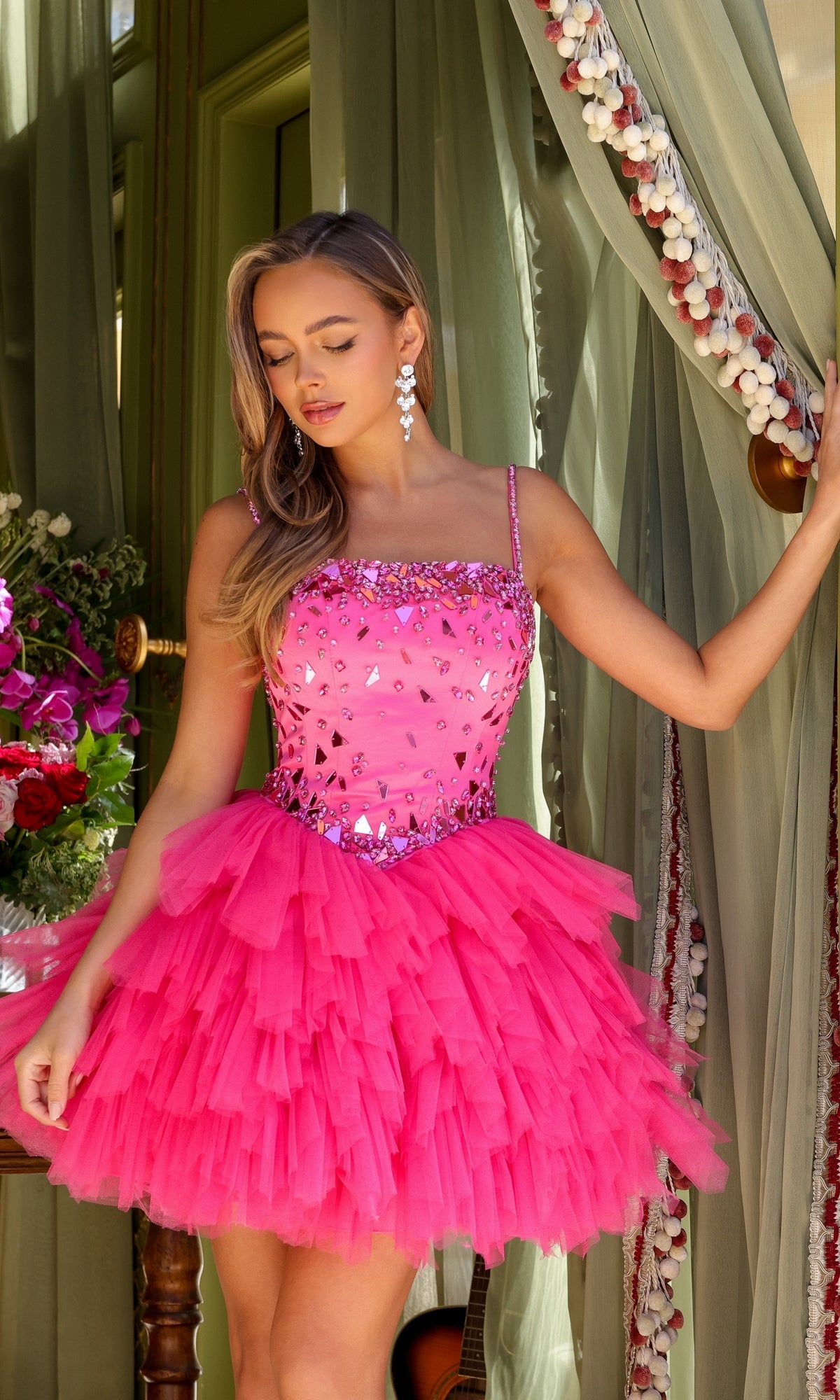A woman in the Ava Presley 29180 Short Tiered Homecoming Dress, a bright pink ruffled tulle style with sequin accents, stands by a green curtain holding it aside, with flowers and a guitar in the background.